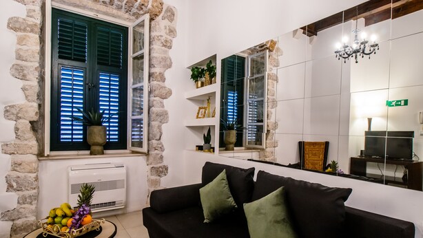 A living room with a black sofa, green cushions, a small table with fruit, and a potted plant. There is a window with shutters, a chandelier, and a television on a stand. - Lovely 1-bed Apartment Niko in Dubrovnik (Dubrovnik)