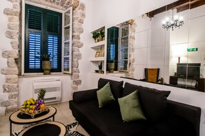 Ein Wohnzimmer mit einer schwarzen Couch, grünen Kissen, einem kleinen Tisch mit Obst und einer Topfpflanze. Es gibt ein Fenster mit Fensterläden, eine Chandelier und einen Fernseher auf einem Schrank.