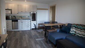 A modern kitchen with white cabinets, a stainless steel refrigerator, and a microwave. There is a dining area with a wooden table and chairs. - West Coast Oceanfront Private Suite with Stunning View (Ucluelet)