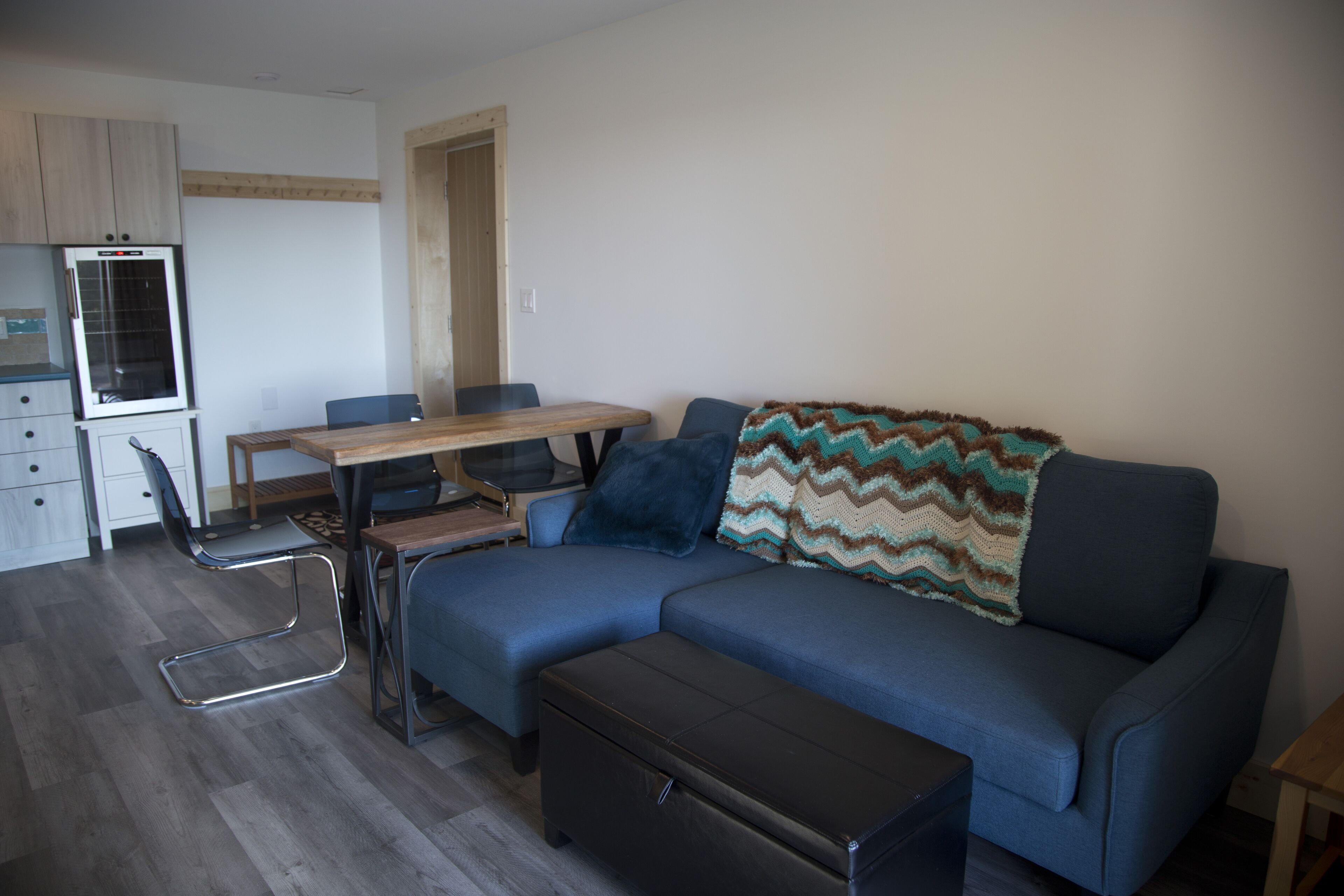Una sala de estar moderna con un sofá azul, una mesa de comedor de madera y una zona de cocina con refrigerador.