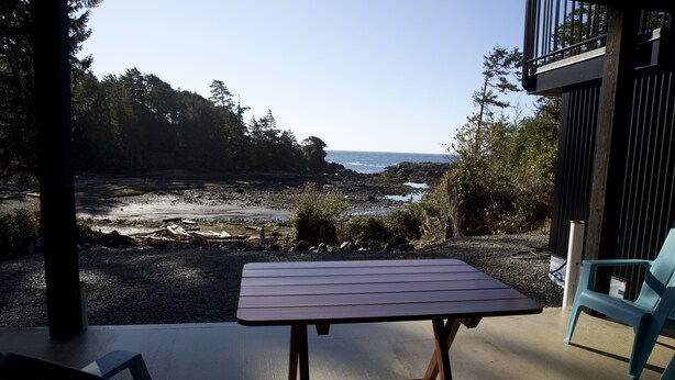 A patio with a table and chairs overlooking a rocky beach and forested area. - West Coast Oceanfront Private Suite with Stunning View (Ucluelet)