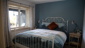 A bedroom with a bed, a window with curtains, a nightstand with a lamp, and a wall with a painting. - West Coast Oceanfront Private Suite with Stunning View (Ucluelet)