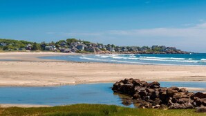 Una playa con casas al fondo y un promontorio rocoso al frente.
