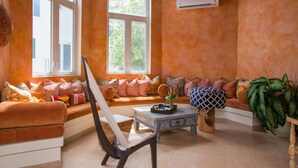 A cozy living room with a sofa, a chair, and a coffee table. There are plants and a window with a view of trees.