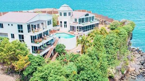A large, multi-story house with a pool, surrounded by lush greenery and a rocky coastline.
