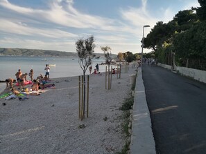Ein Strand mit badenden Menschen und Schwimmern, eine ihn parallel verlaufende Straße und ein ruhiges Meer im Hintergrund.