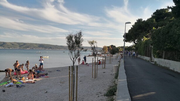 A beach with people sunbathing and swimming, a road parallel to the beach, and a calm sea in the background. - Apartments Vujina - Studio 3 (Kastel Stafilic)