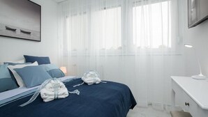 Een moderne slaapkamer met een bed, twee kussens, een nachtkastje, een lamp en een raam met dunne gordijnen.