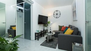 Een moderne woonkamer met een grijze bank, een salontafel en een televisie. Er hangt een klok aan de muur en er staat een plant in de hoek.