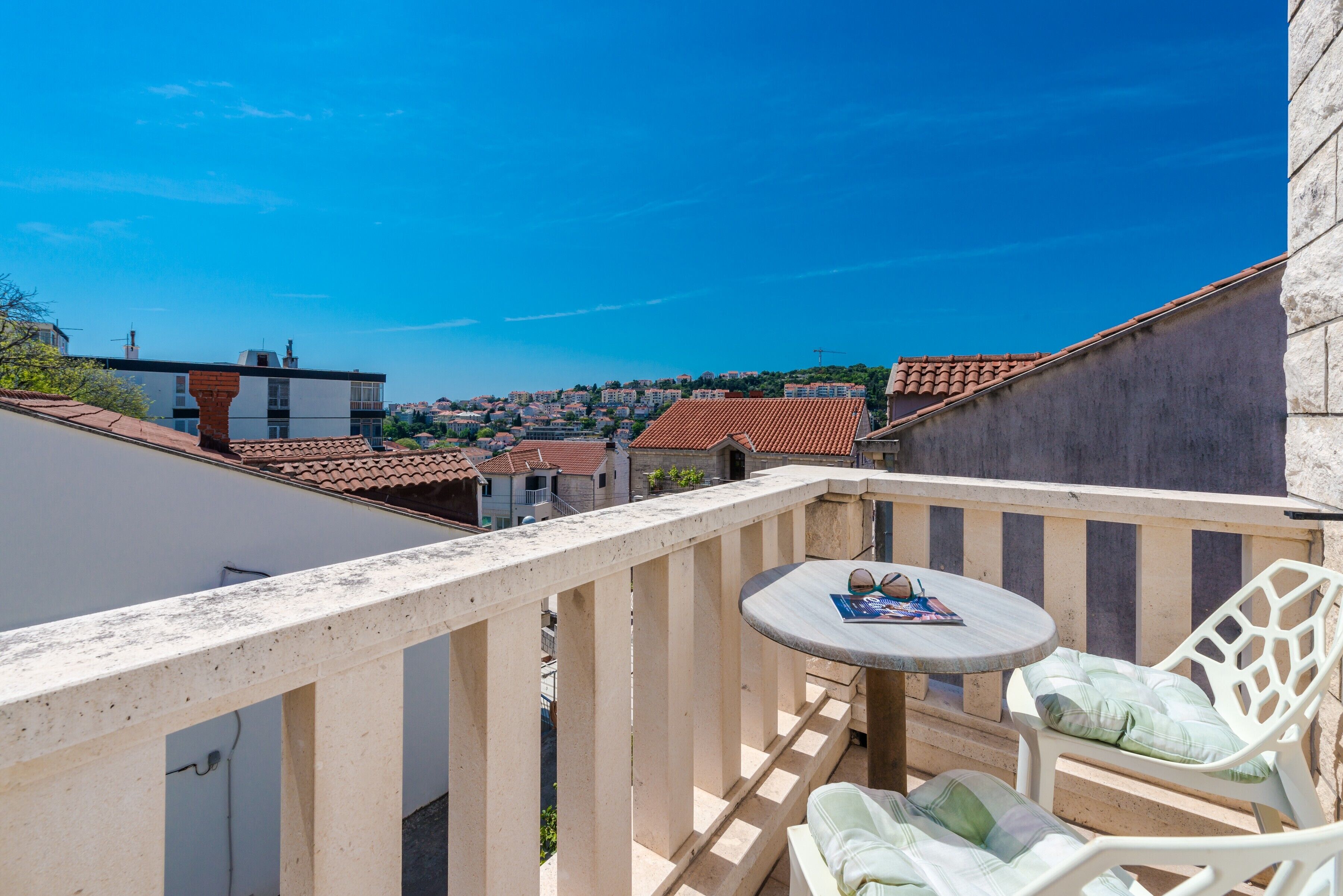 A balcony with a round table and two chairs, overlooking a town with red-tiled roofs.