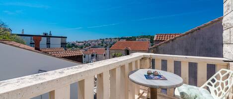 A balcony with a round table and two chairs, overlooking a town with red-tiled roofs.