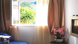Una habitación con cama, ventana con cortinas, una mesita con un florero y un televisor montado en la pared.