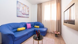 Una sala de estar con un sofá azul, una mesita de centro redonda, un cuadro de veleros, un televisor montado en la pared y una ventana con cortinas.