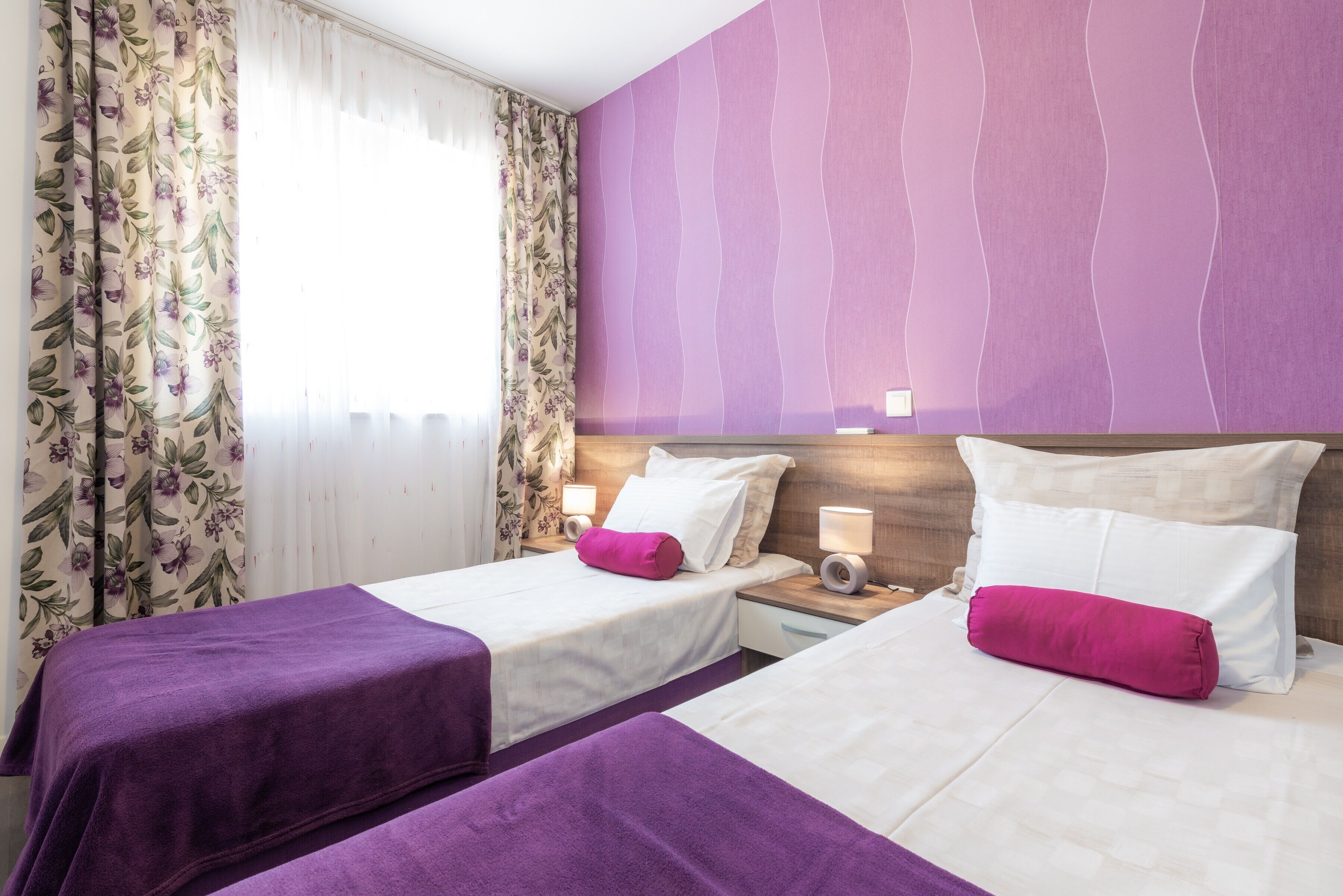 Une chambre d’hôtel avec deux lits, une literie violette, des rideaux à motifs floraux et un mur d’accent violet.