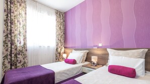 Ein Hotelzimmer mit zwei Betten, violetter Bettwäsche, blumigen Vorhängen und einer violetten Akzentwand.