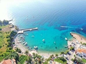 An aerial view of a coastal area with a pier extending into the sea, surrounded by clear blue waters and boats. There are buildings and greenery along the coastline.