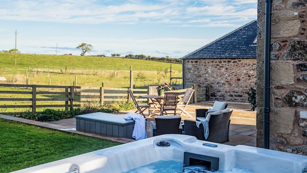 A hot tub with a stone wall and outdoor furniture, surrounded by a grassy field and a stone building. - Step into the Cart House and you will feel instantly at ease. (Duns)
