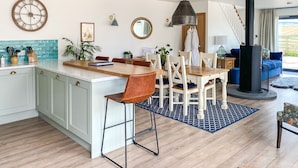 A modern kitchen with a central island, dining area with chairs, a large clock, and a black stove. - Step into the Cart House and you will feel instantly at ease. (Duns)