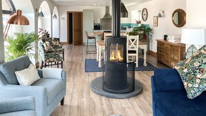 A living room with a central wood-burning stove, a dining area, and a kitchen in the background. - Step into the Cart House and you will feel instantly at ease. (Duns)