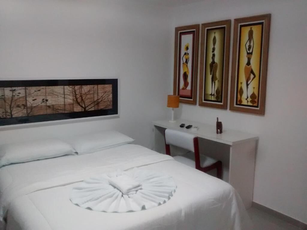 Quarto de hotel com uma cama bem arrumada, uma escrivaninha com abajur e três obras de arte emolduradas na parede.