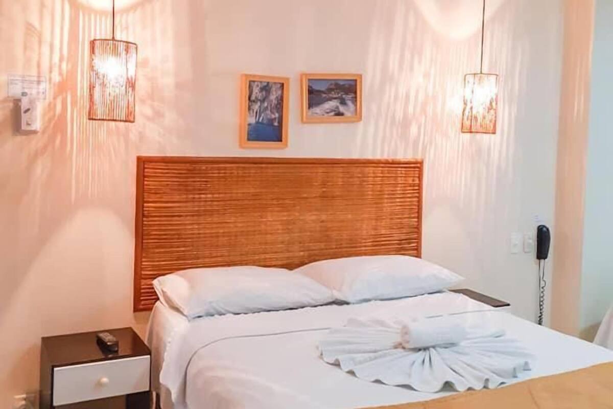 Una cama bien hecha con sábanas blancas y una manta de color marrón, un cabecero de madera y dos lámparas colgantes.