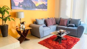 A living room with a grey sofa, a glass coffee table, a red rug, a potted plant, and a painting of a coastal landscape. - HOUSE OF THE SEA, fraction. private with pool, by car 7 minutes from the beach and downtown (Cabo San Lucas)
