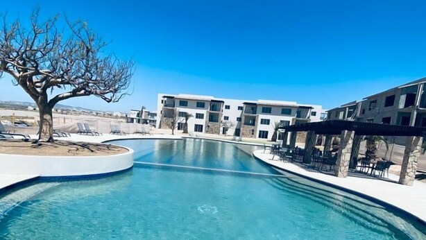 A clear pool with a curved edge, surrounded by a white tiled deck. In the background, there are multi-story buildings with balconies and a clear blue sky. - HOUSE OF THE SEA, fraction. private with pool, by car 7 minutes from the beach and downtown (Cabo San Lucas)