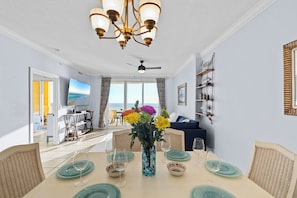 A dining area with a table set for four, a vase with flowers, and a view of the sea through large windows.