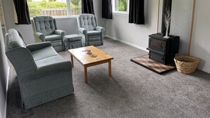 Een woonkamer met grijze banken, een houten salontafel, een zwarte houtkachel en grote ramen met donkergrijze gordijnen.