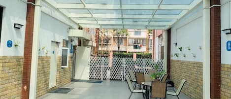 A covered outdoor area with a glass ceiling, a seating arrangement, and a view of a residential building in the background.