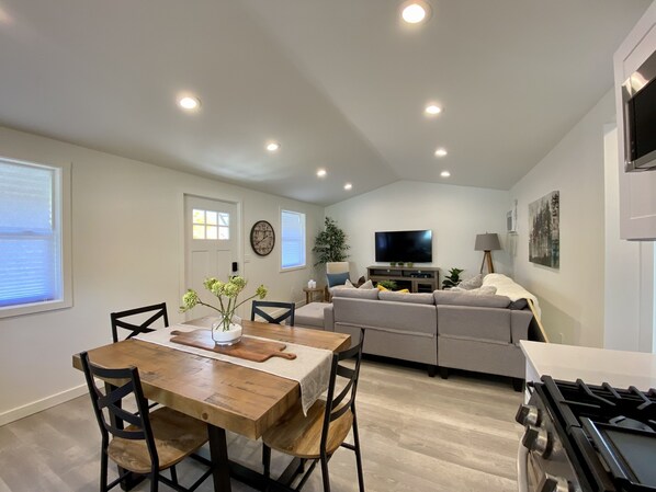A modern kitchen with a stove, dining table, and chairs. There is a living area with a sofa and a flat-screen TV.