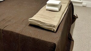 Una cama bien tendida con una colcha marrón estampada, una manta beige y dos toallas blancas dobladas. Se ve una cesta de mimbre blanca apoyada en la pared y una ventana con cristal satinado.