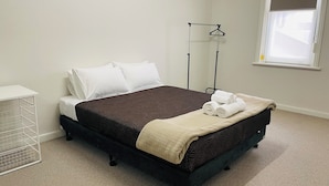 Una cama bien hecha con un cabecero oscuro, almohadas blancas y una manta color beige. Se aprecia una mesita de noche blanca con un estante metálico, una ventana con persiana blanca y una lámpara de pared.