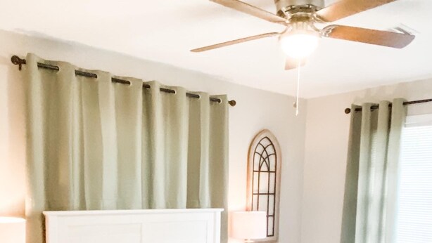 Une chambre avec un grand lit, un ventilateur de plafond et une fenêtre avec des rideaux.