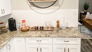 Une cuisine avec des meubles blancs, un plan de travail en granit, une machine à café, un pichet et un plateau contenant des tasses et des verres à café.