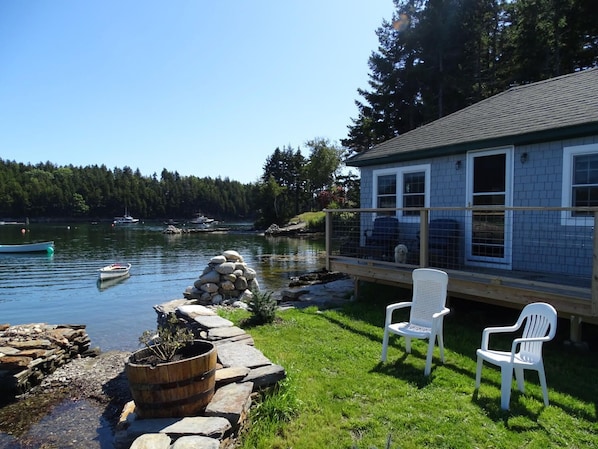 A lakeside house with a deck, a stone wall, and two white chairs.