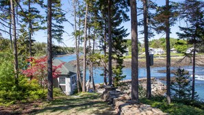 En stenlagd gång leder till en stuga i ett skogsområde, med utsikt över en sjö och hus i bakgrunden.