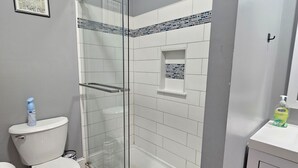 Une salle de bain avec une douche à parois de verre, des carreaux blancs, des toilettes et un lavabo équipé d’un distributeur de savon.