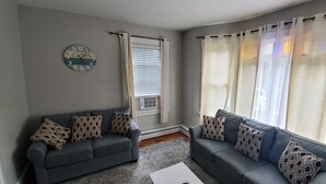 Un salon avec deux canapés gris, des coussins à motifs, une horloge murale ronde et une fenêtre avec des rideaux blancs.