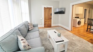 Un salon avec un canapé gris, une table basse blanche, un téléviseur à écran plat fixé au mur et un lave-linge dans la pièce adjacente.