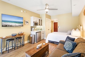 A compact hotel room with a kitchenette, a bed with a white comforter, a wooden coffee table, and a large TV displaying a beach scene.