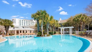 A large, clear swimming pool surrounded by palm trees and lounge chairs, with a hotel building in the background.