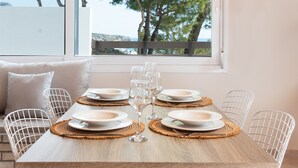 Un coin repas avec une table dressée pour quatre personnes, un luminaire suspendu en fibres tressées et une vue sur des arbres et une étendue d’eau, visible par une fenêtre.