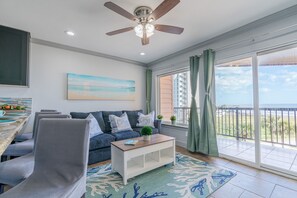 A living room with a sofa, a coffee table, a ceiling fan, and a large window with a view of the beach.