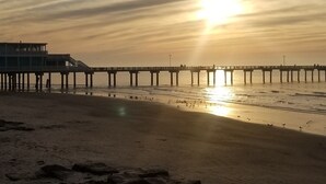 Una spiaggia con un molo che si protende nell'acqua, un tramonto sullo sfondo e rocce sparse sulla sabbia.