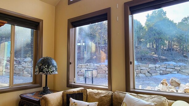 A living room with a beige sofa, two windows with views of a stone wall and trees, and a lamp on a wooden side table. - Private and secluded mountain cabin (Cedar City)