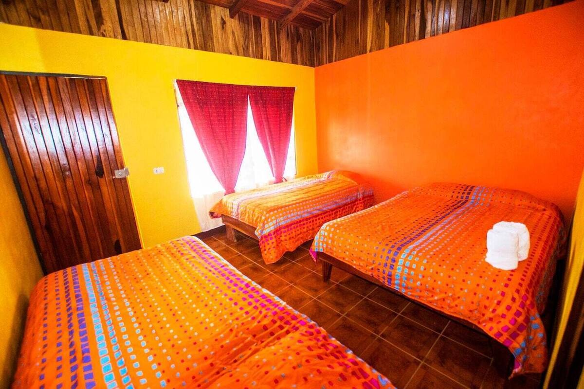 A room with two beds, wooden ceiling beams, and a window with red curtains.