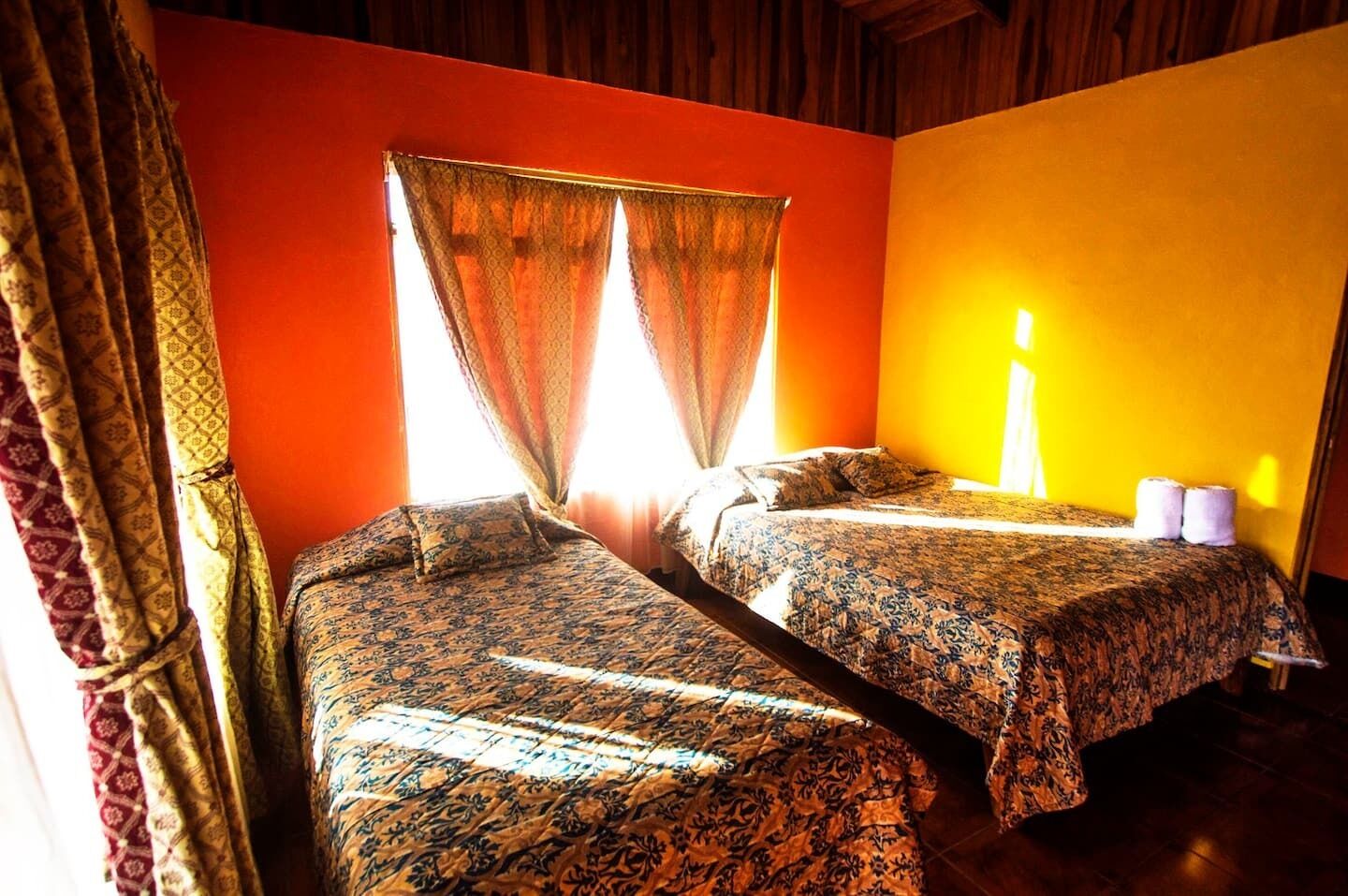 Una habitación con dos camas, una ventana con cortinas y una lámpara de pared.