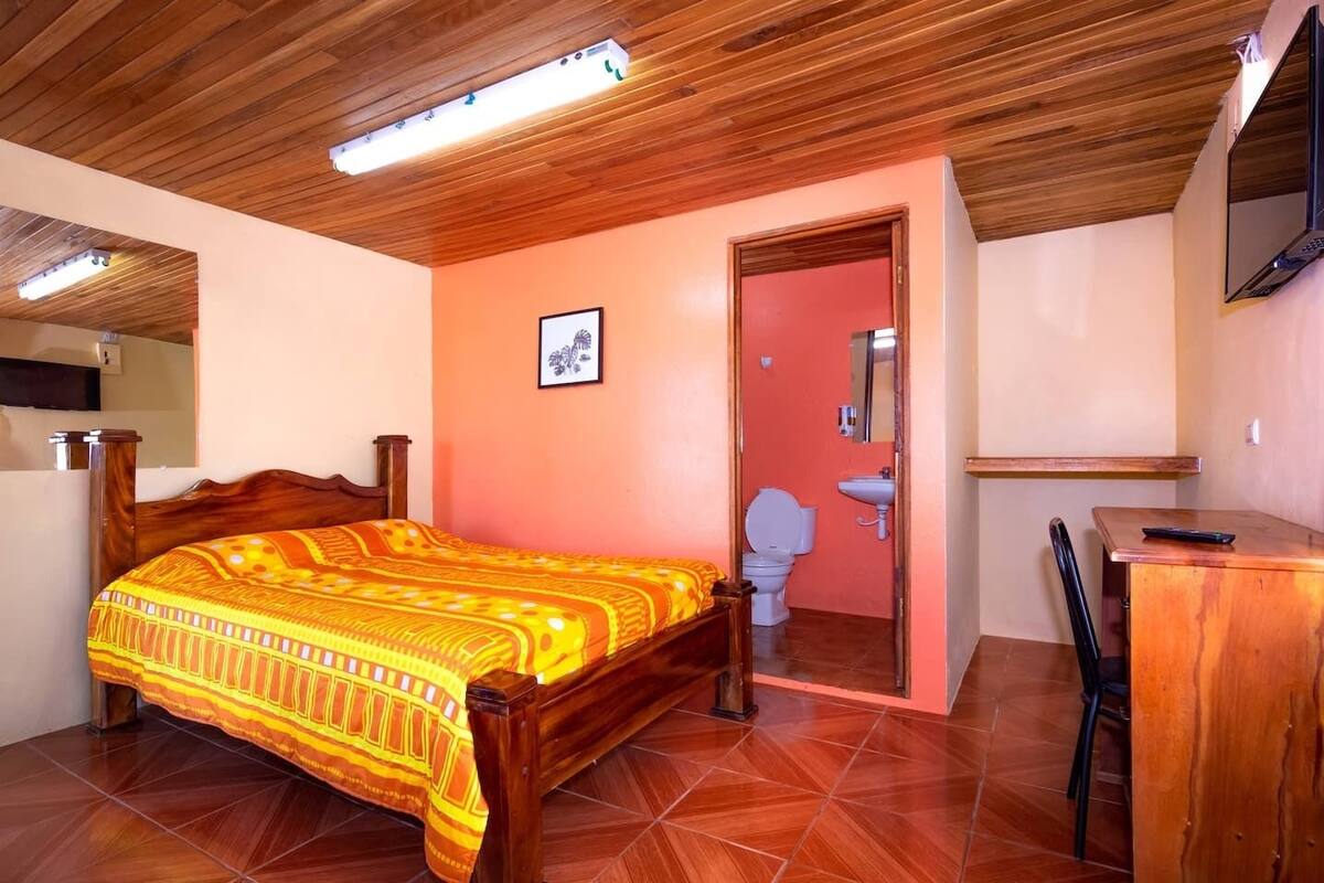 A bedroom with a wooden bed, a desk, a chair, a bathroom with a toilet and sink, and a TV mounted on the wall.