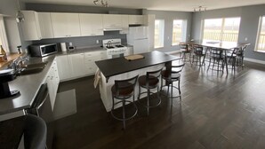 A spacious kitchen with white cabinets, a central island with stools, a dining area with a table and chairs, and large windows offering a view. - Red Roof Chalet in close proximity to beaches, trails, golf and Blue Mountains. (Meaford)